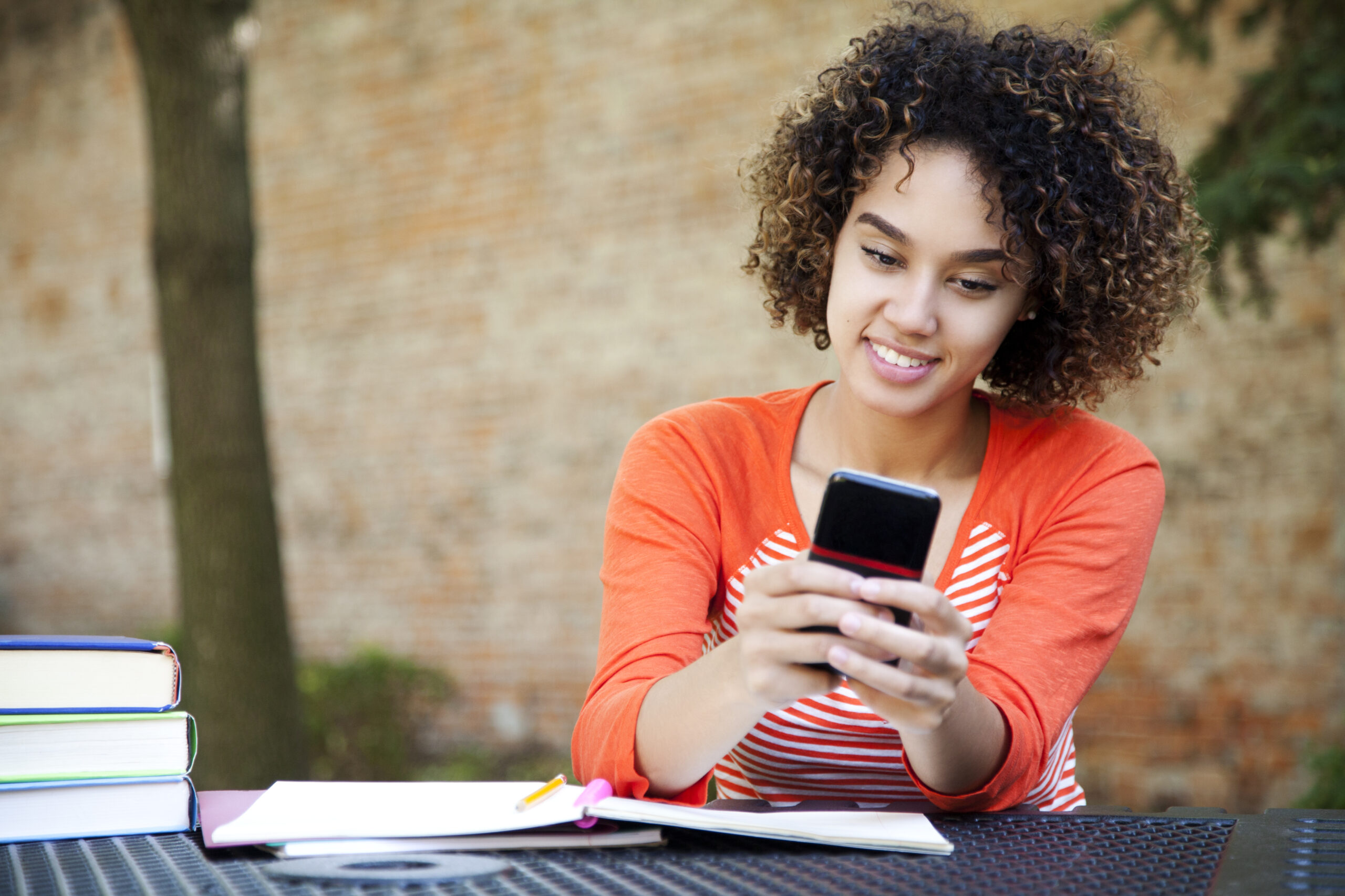 Ethnic female student with curly hair wearing wearing an orange shirt and texting with her smart phone